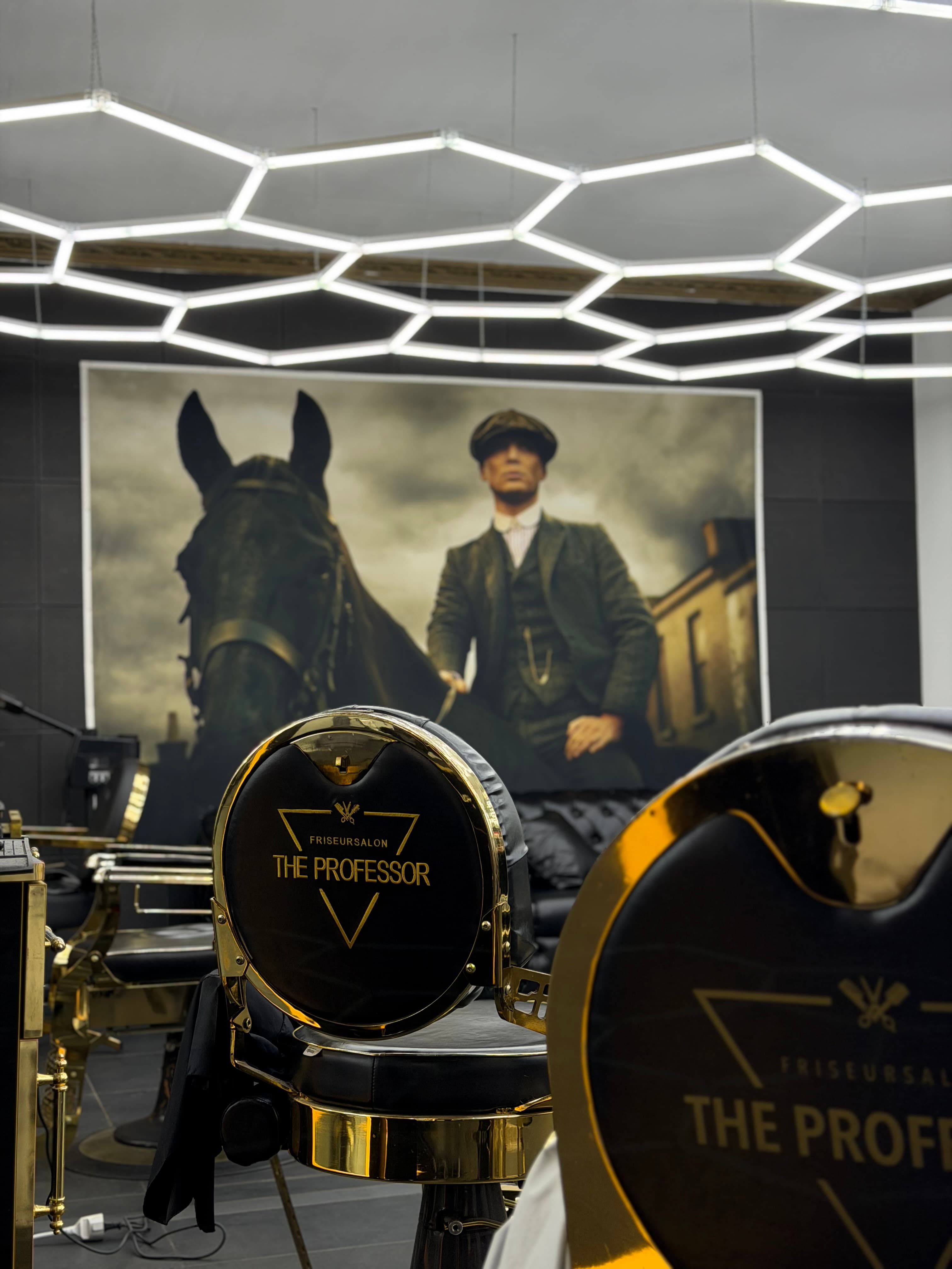 The Professor Barbershop in Kirchheim unter Teck — markenbedruckte Barbierstühle vor Peaky-Blinders-Wandbild und Hexagon-LED-Beleuchtung